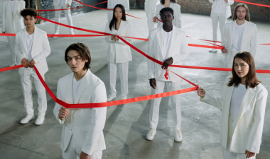 Various people stand in a room holding tape in a pattern that shows connectedness.