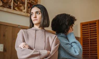 Two women stand back-to-back indoors, appearing upset.