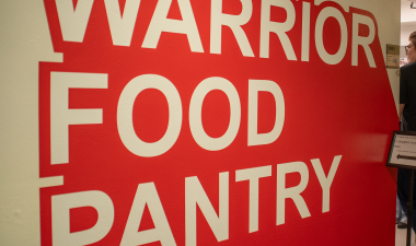 Bold red wall with the words "Warrior Food Pantry" in large white letters. A sign and a person are visible to the right, suggesting entrance.
