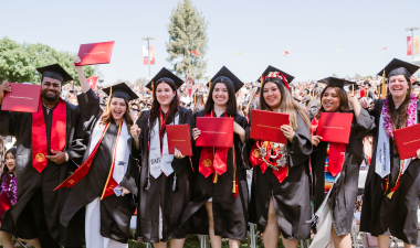 A group of seven graduates in caps and gowns joyfully hold diplomas, celebrating on a sunny day outdoors, surrounded by a large, cheering crowd.