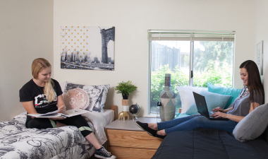 Two women relax in a bright dorm room. One reads on the left bed; the other uses a laptop on the right. The scene is casual and studious.