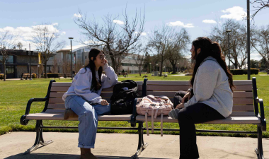 Two people sit and talk on a park bench under a sunny sky, with backpacks and bare trees around, conveying a relaxed and casual atmosphere.