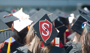 Graduates wearing black caps and gowns are seated at a ceremony. One cap features a red and white emblem. The scene conveys a celebratory mood.