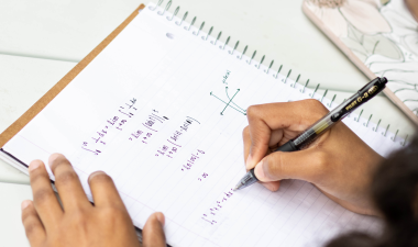 A person is writing math equations in a spiral notebook on a white desk. Their hands are focused and engaged. An artistic phone case is visible nearby.