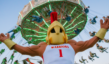 Titus stands with arms wide open in front of a colorful swing ride. People are joyfully swinging high in the air.