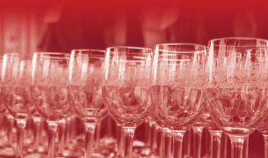 Rows of empty wine glasses with elegant script etched onto them, arranged closely together under a soft red gradient filter, creating a refined and festive atmosphere.