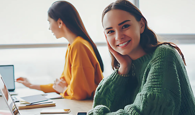 A young woman sits a desk and faces the camera with a smile on her face.