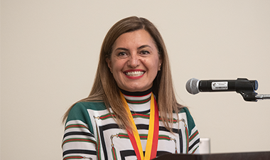 A woman smiling warmly stands at a podium with a microphone. She wears a colorful, striped top and a yellow and red lanyard, against a plain background.