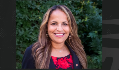 Smiling woman with long hair, wearing a black blazer and red patterned top against a lush green leafy background. The mood is warm and professional.