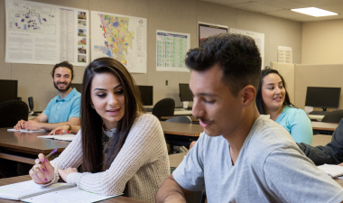 A diverse group of students sits in a classroom, engaged in discussion. Papers and computers are on the desks, creating a collaborative and focused atmosphere.