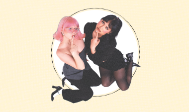 Two people kneeling, one with pink hair, glasses, and a playful expression, the other with dark hair and black outfit, both posing thoughtfully.