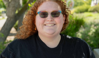 Smiling person with curly red hair and sunglasses wears a black shirt reading "WARRIOR Strong" in red. Trees and greenery form the sunny background.