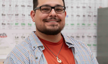 A man with glasses and a beard, smiling, sits at a lab table with a microscope. He wears a red shirt and checkered jacket, conveying a friendly atmosphere.