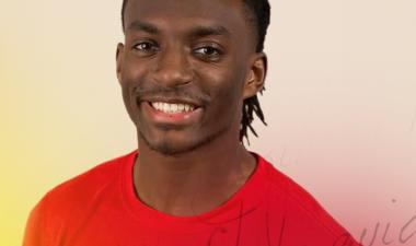 Smiling man wearing a red shirt, with short dreadlocks. The background is softly blurred in shades of light beige and yellow, conveying a cheerful tone.