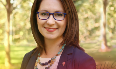 A woman with shoulder-length brown hair and glasses smiles warmly. She's wearing a colorful beaded necklace and a dark blazer, set against a sunlit, leafy background. The tone is professional and approachable.