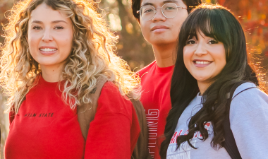 Three Stanislaus State students wearing campus apparel pose outdoors with autumn foliage in the background.