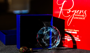 A crystal Rogers Scholars commemorative award displayed in front of a blue presentation box, with two gold pins and a red Rogers Scholars Luncheon program in the background.