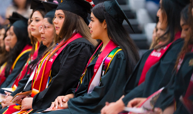 Stanislaus State graduates wearing caps, gowns and colorful stoles sit together during the Chicanx/Latine Graduation Celebration as family and friends look on.