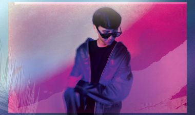 A young person in dark sunglasses and a blue jacket sits with arms folded against a pink and purple backdrop, conveying a cool, introspective mood.