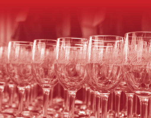 Rows of empty wine glasses with elegant script etched onto them, arranged closely together under a soft red gradient filter, creating a refined and festive atmosphere.