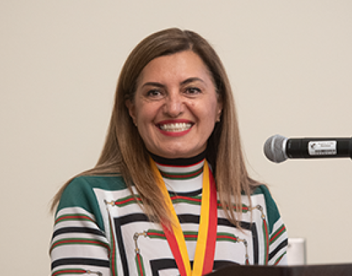 A woman smiling warmly stands at a podium with a microphone. She wears a colorful, striped top and a yellow and red lanyard, against a plain background.
