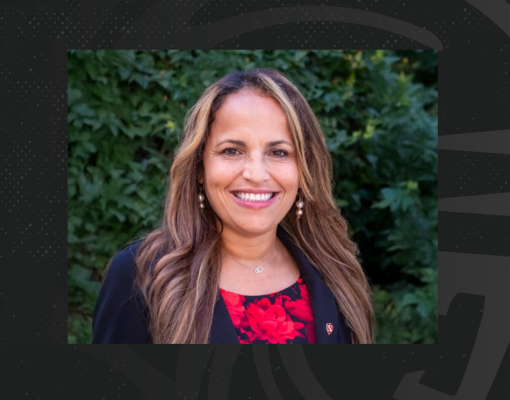 Smiling woman with long hair, wearing a black blazer and red patterned top against a lush green leafy background. The mood is warm and professional.