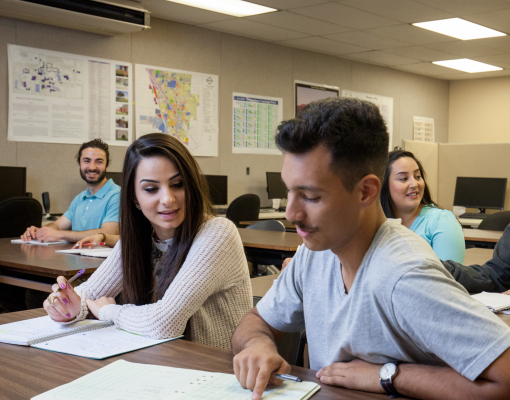 A diverse group of students sits in a classroom, engaged in discussion. Papers and computers are on the desks, creating a collaborative and focused atmosphere.