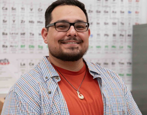 A man with glasses and a beard, smiling, sits at a lab table with a microscope. He wears a red shirt and checkered jacket, conveying a friendly atmosphere.