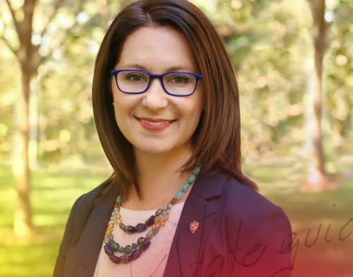 A woman with shoulder-length brown hair and glasses smiles warmly. She's wearing a colorful beaded necklace and a dark blazer, set against a sunlit, leafy background. The tone is professional and approachable.
