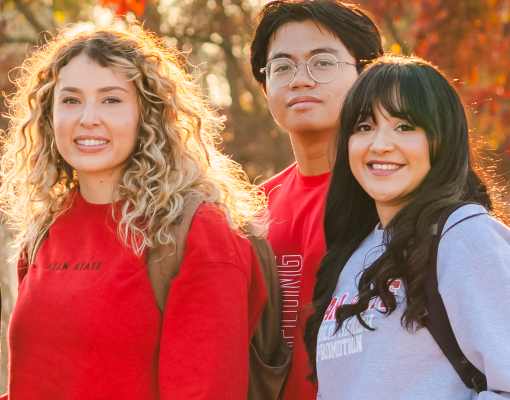 Three Stanislaus State students wearing campus apparel pose outdoors with autumn foliage in the background.