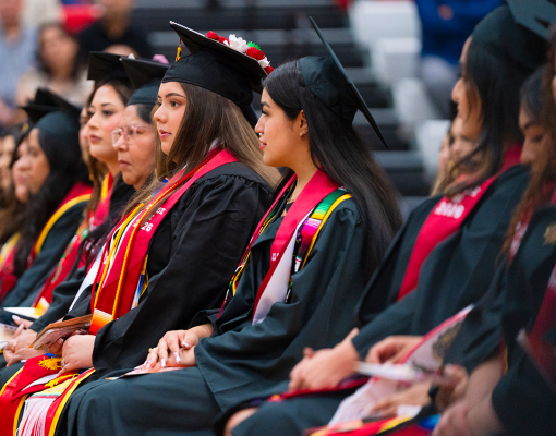 Stanislaus State graduates wearing caps, gowns and colorful stoles sit together during the Chicanx/Latine Graduation Celebration as family and friends look on.