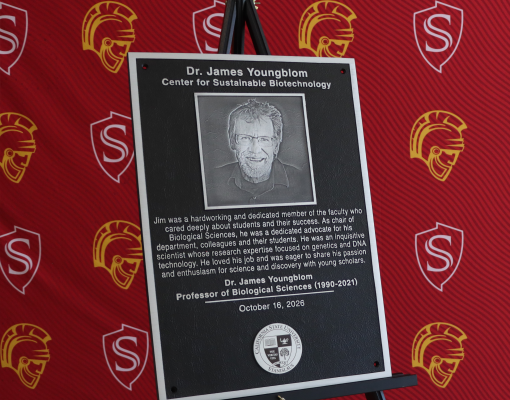 A commemorative plaque honoring James Youngblom, featuring his portrait and dedication to students, teaching and scientific research, stands on an easel against a red Stanislaus State backdrop.