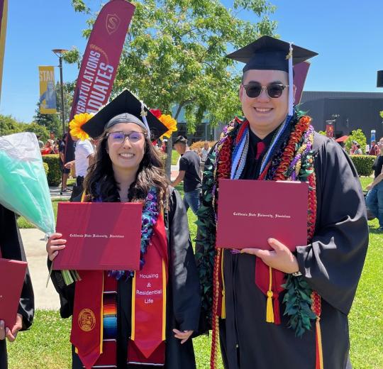 male and female student in grad regalia holding diplomas and smiling