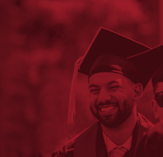 Two graduates in caps and gowns, one wearing sunglasses, celebrate during a ceremony in a vibrant red-toned background.