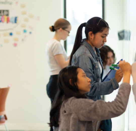 Four students are writing on a whiteboard wall.