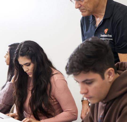 A teacher in a dark shirt with a school logo stands over a student in a pink sweater focused on her work. Other students study nearby, creating a focused, studious environment.