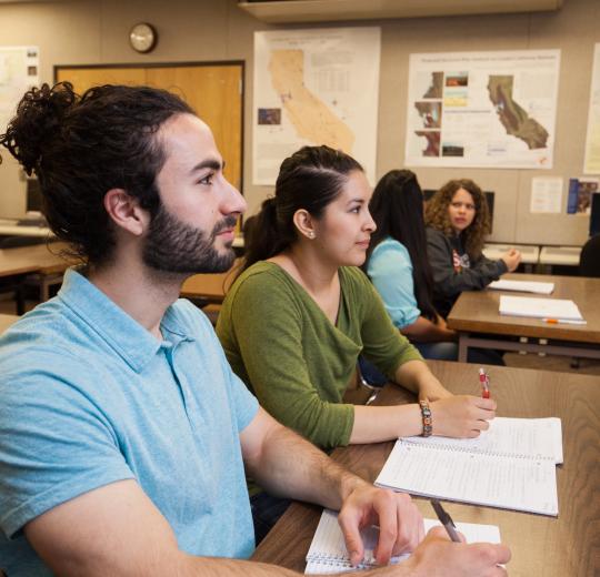 Students sit attentively in a classroom, taking notes. Maps of California adorn the walls, creating an educational and focused atmosphere.