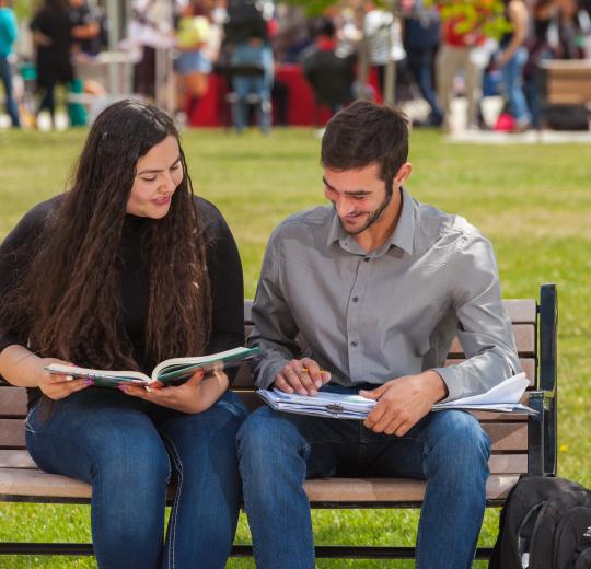A young woman and man sit closely on a wooden bench in a park, studying textbooks and smiling. Around them, blurred crowds suggest a lively campus setting.