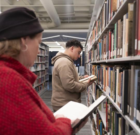 People browsing books in a library aisle, one wearing a red coat and black hat, another in a brown hoodie, both engrossed in reading.