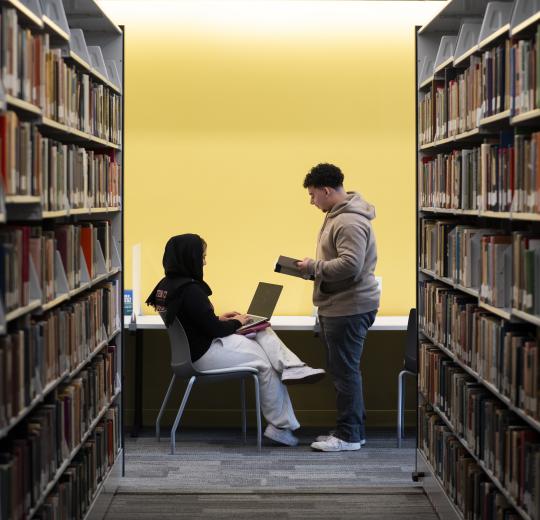 A student sits at a library desk with a laptop, while another stands holding a book. Surrounded by shelves, they engage in focused discussion.