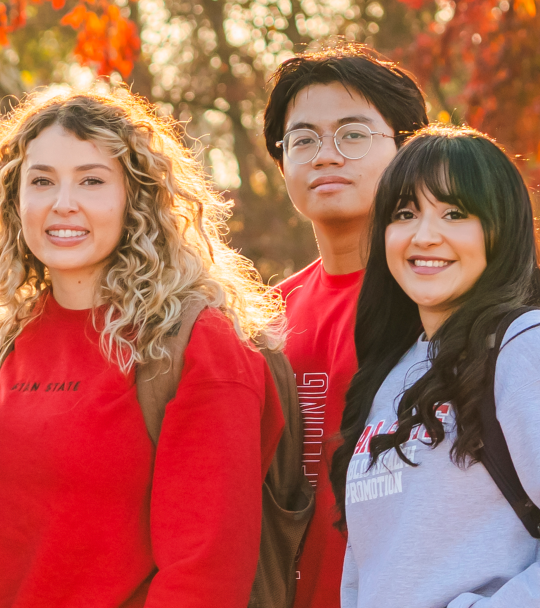 Three Stanislaus State students wearing campus apparel pose outdoors with autumn foliage in the background.