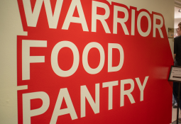 Bold red wall with the words "Warrior Food Pantry" in large white letters. A sign and a person are visible to the right, suggesting entrance.
