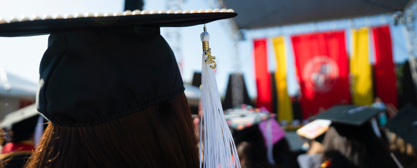 Close-up of a graduate wearing a decorated cap with a white tassel, facing a stage adorned with vibrant banners. The scene conveys achievement and celebration.